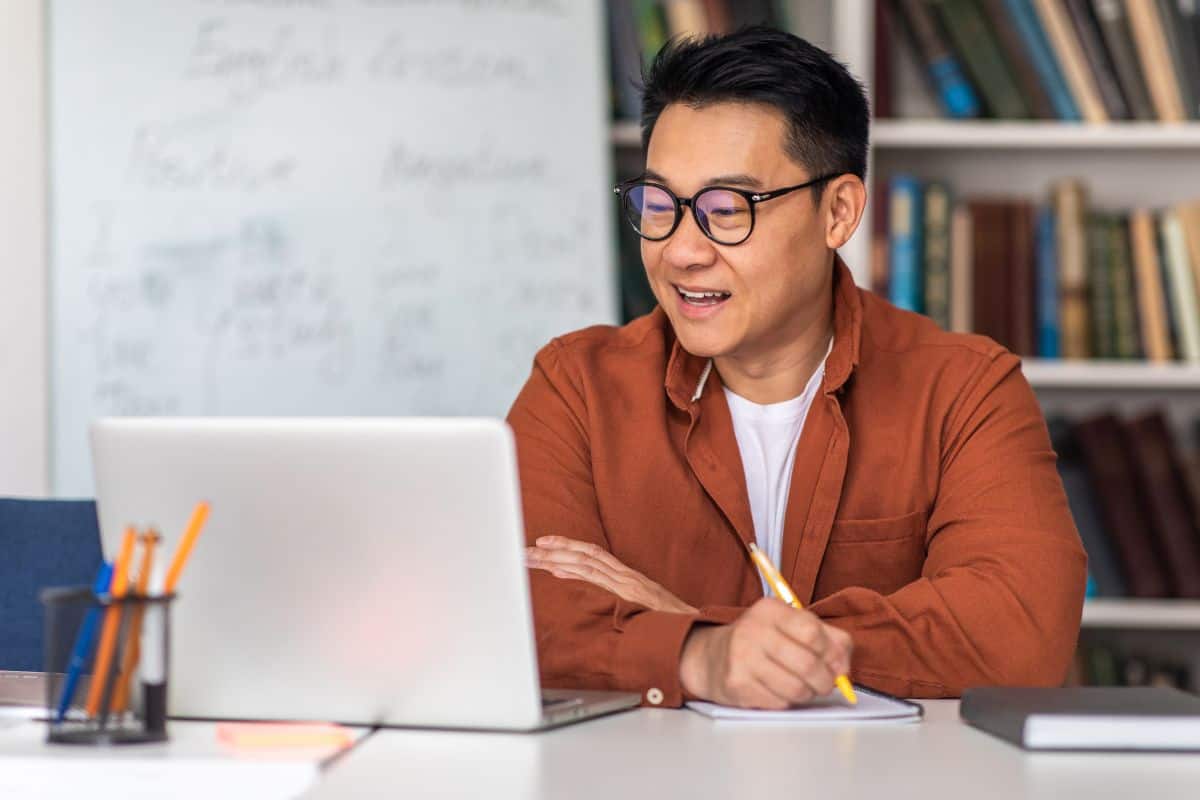 Asian tutor having online class via laptop taking notes indoor
