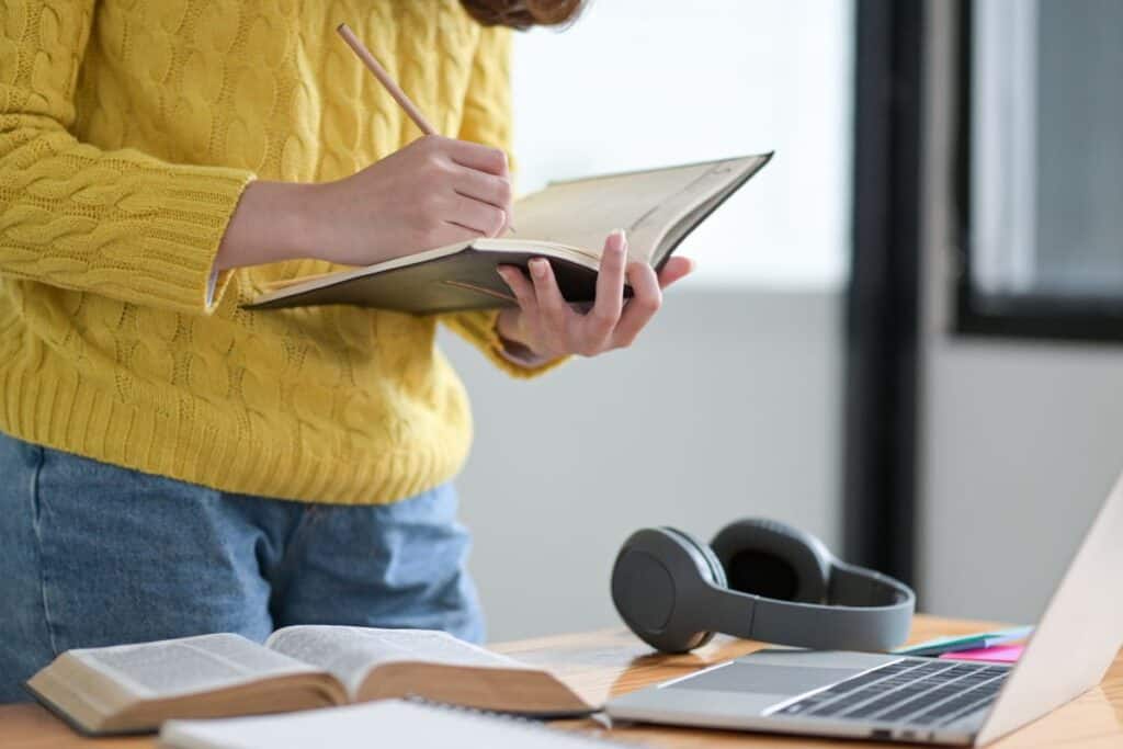 A female student takes notes in a notebook during an online class on a laptop