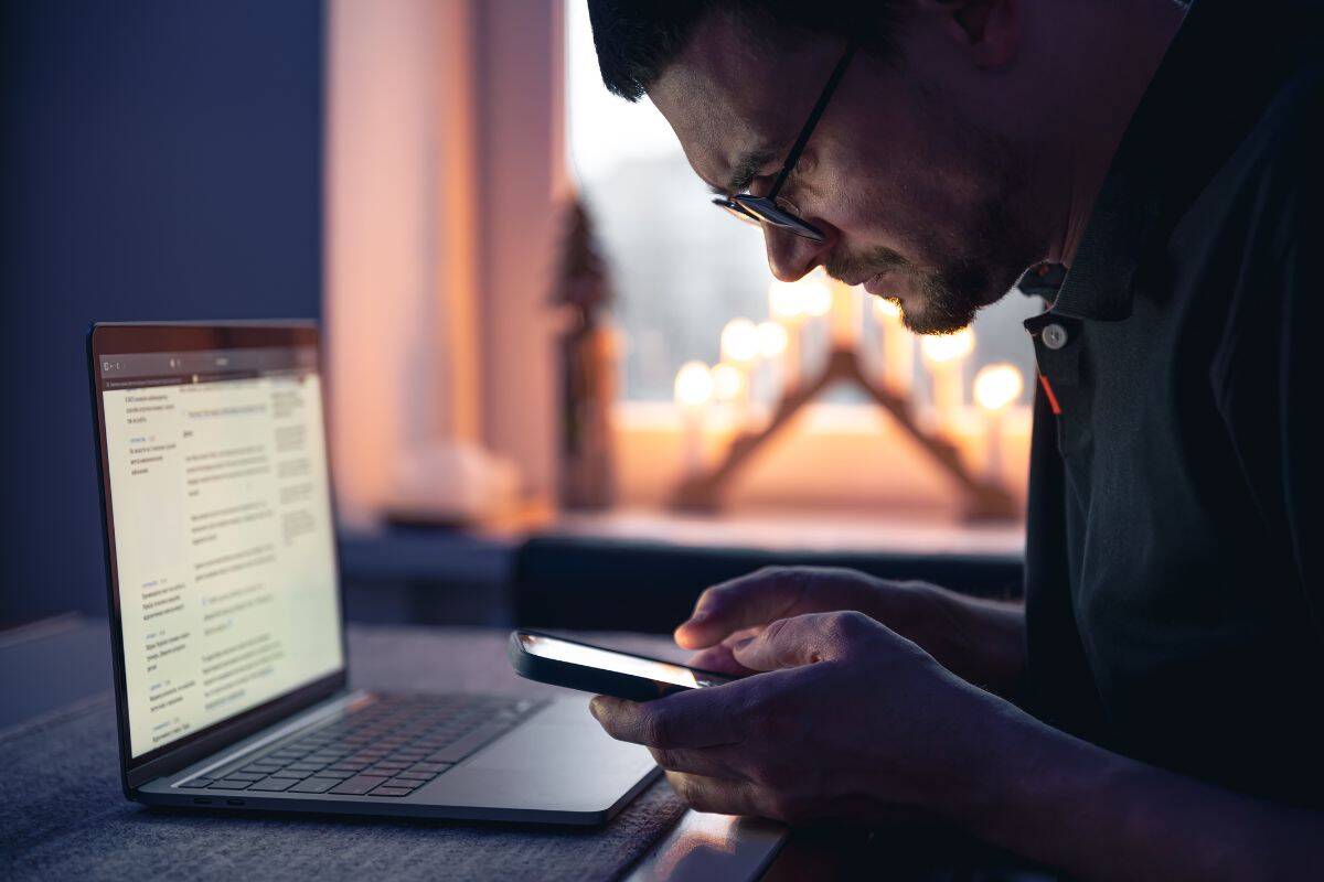 A man with a smartphone sits in front of a laptop late at night
