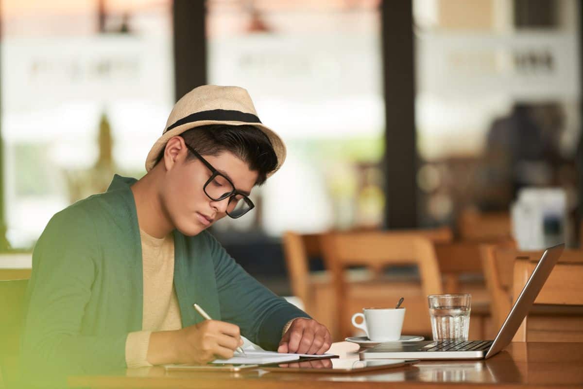 Homem asiático jovem e elegante sentado em um café com laptop e escrevendo em um caderno