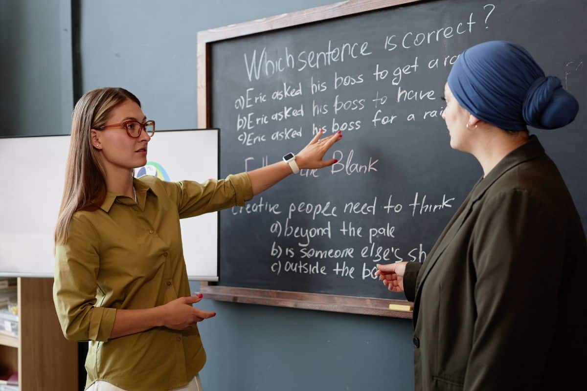 Young female english teacher assisting student standing at blackboard