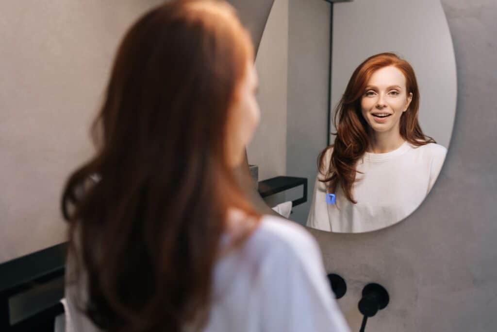 Cheerful young woman smiling to reflection in mirror standing