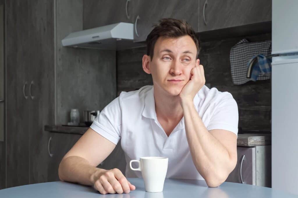 Sleepy man naps at table and drinks cup of coffee in kitchen