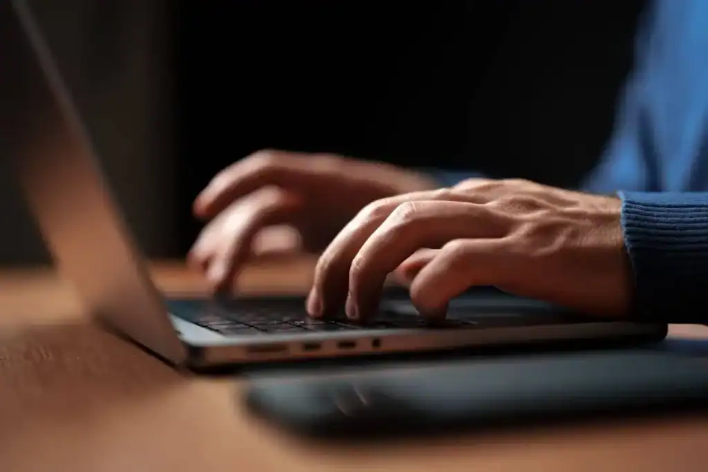 Mani che digitano su un computer portatile in uno spazio di lavoro poco illuminato nelle ore serali