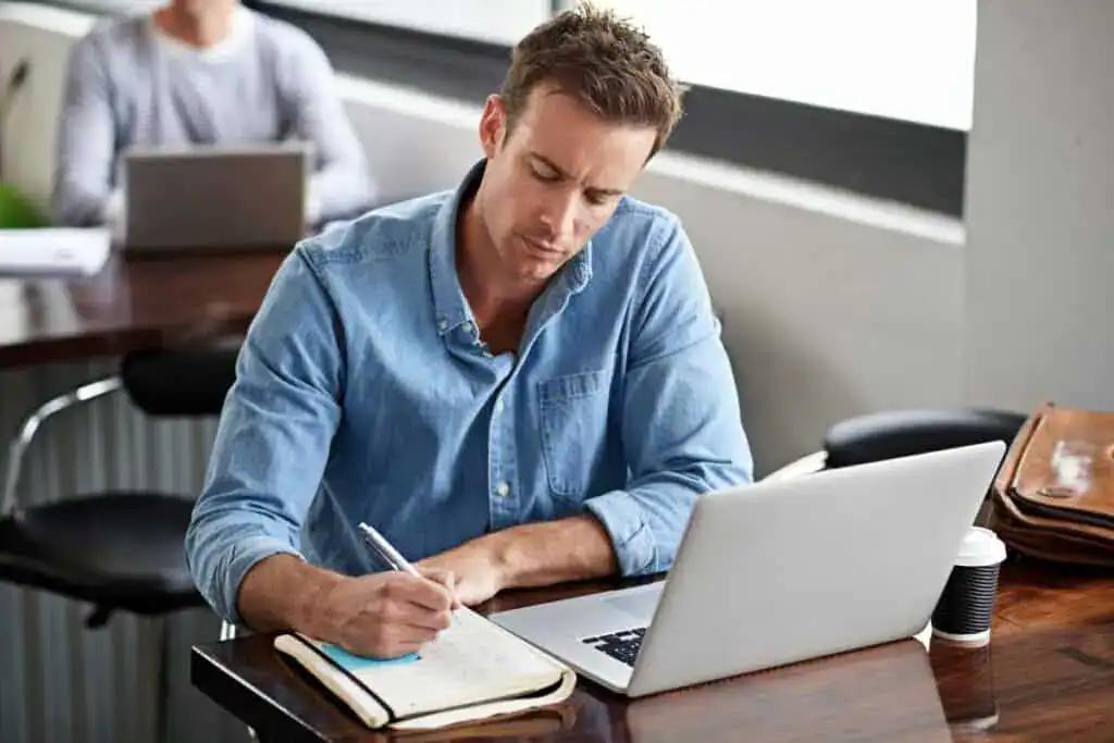 Man writing on book and laptop in modern office for information with project